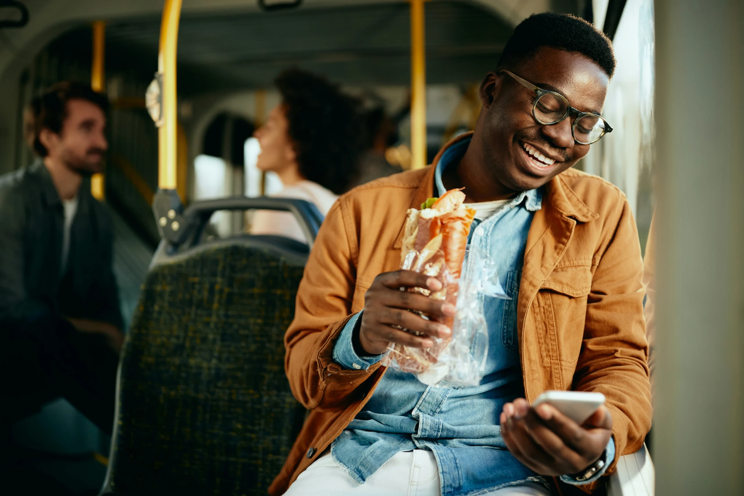 happy black man eating sandwich using smart phone while commuting by bus